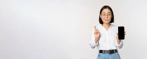 Mujer asiática joven esperanzada, mostrando la pantalla del teléfono móvil, interfaz de la aplicación y los dedos cruzados, esperando smth, pidiendo deseo o pagando, de pie sobre fondo blanco — Foto de Stock