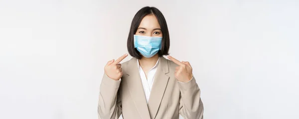 Koncepcja koronawirusa i ludzi biznesu. Azjatka przedsiębiorca wskazując palcami na medyczną maskę twarzy w miejscu pracy, stojąc na białym tle — Zdjęcie stockowe