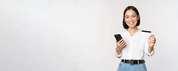 Concepto de compras online. Sonriendo chica asiática moderna muestra su tarjeta de crédito, sostiene el teléfono móvil, orden con el teléfono inteligente, de pie sobre fondo blanco — Foto de Stock