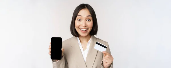 Företagskvinna med glad, entusiastisk ansikte, visar kreditkort och smartphone app-skärm, står i kostym över vit bakgrund — Stockfoto