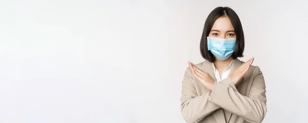 Concepto de Coronavirus y lugar de trabajo. Imagen de mujer de negocios asiática, mujer en máscara facial médica muestra parada, gesto de prohibición cruzada, se para sobre fondo blanco —  Fotos de Stock