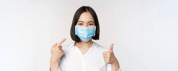 Porträtt av asiatisk flicka i medicinsk mask visar tummen upp skylt och pekar på hennes covid skydd, står över vit bakgrund — Stockfoto