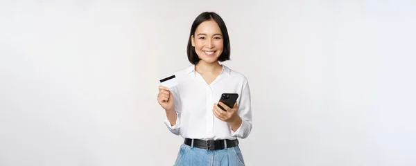 Concepto de compras online. Imagen de una joven asiática moderna sosteniendo tarjeta de crédito y teléfono inteligente, comprando con aplicación de teléfono inteligente, pagando sin contacto, de pie sobre fondo blanco — Foto de Stock