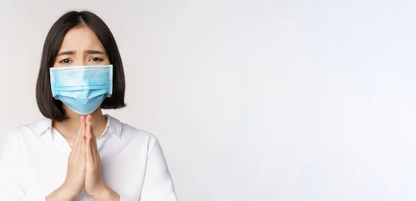 Portrait de femme asiatique dans le masque facial médical de covid, suppliant, demandant de l'aide, dire s'il vous plaît, debout sur fond blanc — Photo