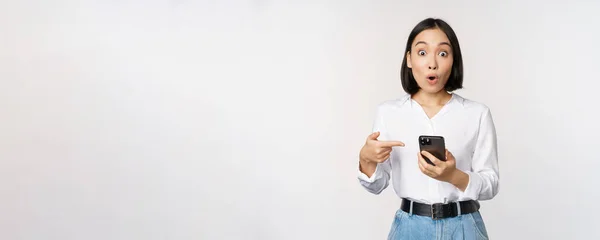 Menina incrível falando sobre o aplicativo do smartphone, apontando para o telefone enquanto olha impressionado com a câmera, de pé contra o fundo branco — Fotografia de Stock