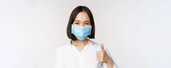 Covid ve sağlık hizmeti konsepti. Tıbbi yüz maskesi takan Asyalı bir kadının portresi ve beyaz arka planda duran, baş parmağını kaldıran, — Stok fotoğraf