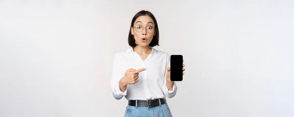 Захоплена молода азіатка, вказуючи пальцем на екран смартфона, показує рекламу на мобільному телефоні, білому фоні — стокове фото
