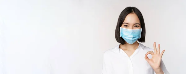 Ковид и концепция здоровья. Портрет азиатской женщины в медицинской маске и с нормальным знаком, стоящей на белом фоне — стоковое фото