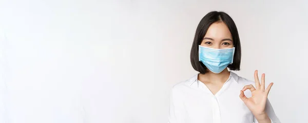 Covid e conceito de saúde. retrato de ásia mulher vestindo médico rosto máscara e mostrando ok sinal de pé sobre branco fundo — Fotografia de Stock