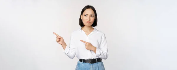 Besviken, ledsen ung asiatisk kvinna pekar och tittar vänster med upprörd, sura ansiktsuttryck, står över vit bakgrund — Stockfoto