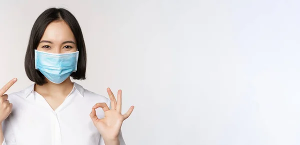 Retrato de chica asiática en máscara médica mostrando signo de bien y apuntando a su protección covid, de pie sobre fondo blanco —  Fotos de Stock