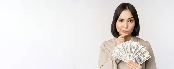 Bild av asiatisk företagskvinna, glad affärskvinna visar pengar, kontanter dollar och tänkande, står i kostym över vit bakgrund — Stockfoto