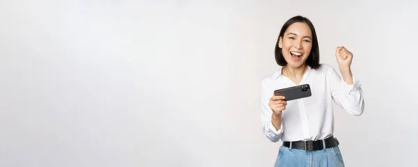 Happy asian woman do winner dance, triumphing, winning on mobile phone video game, holding smartphone horizontal position and celebrating, white background — Stock Photo, Image
