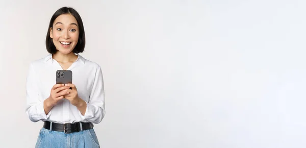 Excited asian woman smiling, reacting to info on mobile phone, holding smartphone and looking happy at camera, standing over white background — Stock Photo, Image