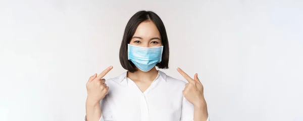 Bild av ung asiatisk kvinna som pekar på sig själv när hon bär medicinsk ansiktsmask, begreppet covid-19 skydd, stående över vit bakgrund — Stockfoto