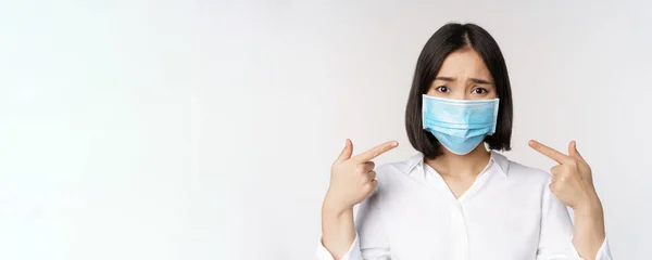 Close up de triste menina asiática em máscara facial médica apontando para a cabeça e olhando chateado, de pé sobre fundo branco — Fotografia de Stock