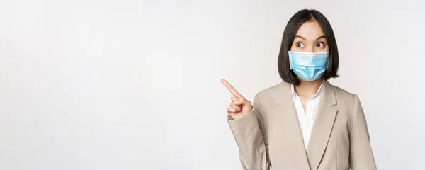 Empresária entusiasmada apontando os dedos à esquerda, vestindo máscara médica da pandemia covid-19, em pé sobre fundo branco — Fotografia de Stock
