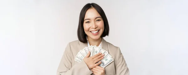 Šťastná asijská podnikatelka držící hotovost, objímající dolary peníze a usmívající se, stojící nad bílým pozadím v obleku — Stock fotografie