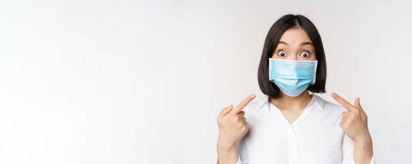 Obraz zdumionej młodej azjatki wskazującej na siebie w medycznej masce na twarzy, stojącej nad białym tłem — Zdjęcie stockowe