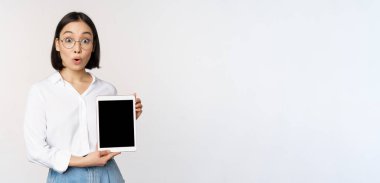 Hevesli Asyalı kadın, gözlüklü ofis çalışanı dijital tablet ekranı gösteriyor, cihaz ekranında bilgi gösteriyor, beyaz arka planda duruyor.