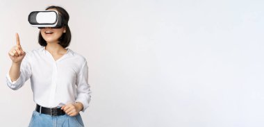 VR kulaklık kullanan Asyalı genç bir kadın. Sanal gerçeklik gözlüklü Koreli kız güler yüzlü, beyaz arkaplanı işaret ediyor.