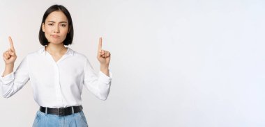 Şüpheci Asyalı kadının suratını buruşturması, suratını asması, parmaklarını kaldırıp reklamını göstermesi, beyaz arka planda durması.