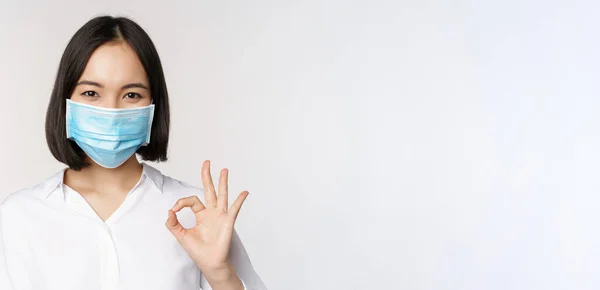 Concepto de covidio y salud. Retrato de una mujer asiática usando mascarilla médica y mostrando un signo aceptable, de pie sobre fondo blanco —  Fotos de Stock