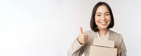 Sorrindo asiático empresária, mostrando polegares para cima e caixas com entrega bens, preparar ordem para o cliente, de pé sobre fundo branco — Fotografia de Stock