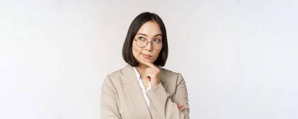 Портрет вдумливої азіатської бізнес-леді в окулярах, роблячи припущення, думаючи, стоячи в бежевому костюмі на білому тлі — стокове фото