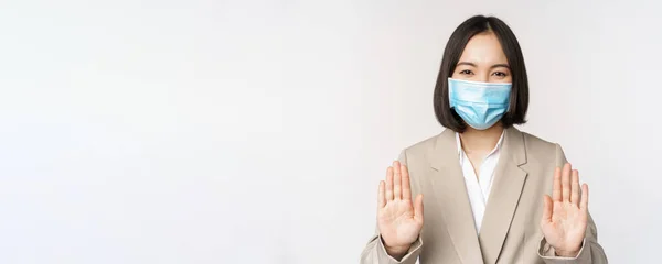 Coronavirus y concepto de trabajo. Retrato de mujer asiática de oficina, mujer en el lugar de trabajo con mascarilla médica y mostrando stop, gesto de prohibición, fondo blanco —  Fotos de Stock