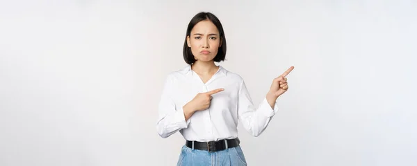 Sorglig och dyster asiatisk kvinna ser besviken, klagar på banner eller reklam, pekar finger rätt på promo och rynka pannan upprörd, vit bakgrund — Stockfoto