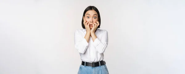Portrait of scared asian woman watching smth scary, biting fingers on hands and looking at camera frightened, standing over white background — Stock Photo, Image