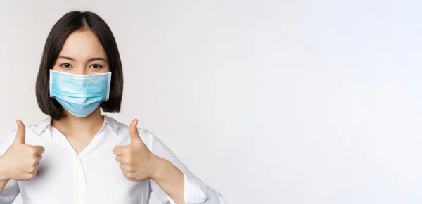 Retrato de sonriente asiática oficina dama en médico mascarilla, mostrando los pulgares hacia arriba, recomendando smth, de pie sobre fondo blanco —  Fotos de Stock