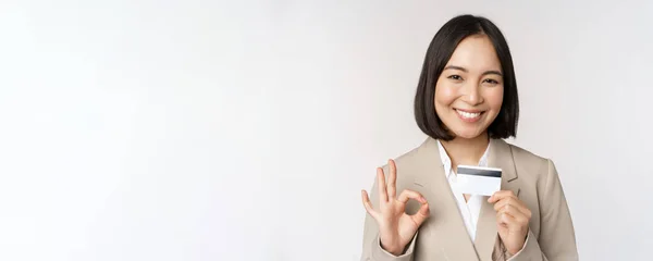 Secretario de oficina sonriente, mujer corporativa asiática mostrando tarjeta de crédito y signo de aprobación, recomendando banco, de pie sobre fondo blanco en traje beige — Foto de Stock