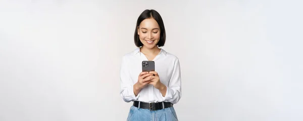 Elegante chica asiática moderna utilizando la aplicación de teléfono móvil, charlando en el teléfono celular y sonriendo, de pie en blusa blanca contra el fondo del estudio — Foto de Stock