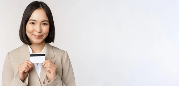 Smiling office clerk, mujer corporativa asiática mostrando tarjeta de crédito, de pie sobre fondo blanco en traje beige — Foto de Stock
