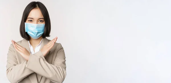 Concepto de Coronavirus y lugar de trabajo. Imagen de mujer de negocios asiática, mujer en máscara facial médica muestra parada, gesto de prohibición cruzada, se para sobre fondo blanco —  Fotos de Stock