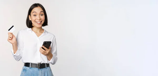 Concepto de compras online. Imagen de una joven asiática moderna sosteniendo tarjeta de crédito y teléfono inteligente, comprando con aplicación de teléfono inteligente, pagando sin contacto, de pie sobre fondo blanco — Foto de Stock