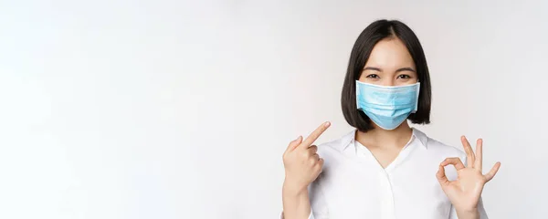 Портрет азиатской девушки в медицинской маске показывает знак одобрения и указывает на ее ковидовую защиту, стоя на белом фоне — стоковое фото