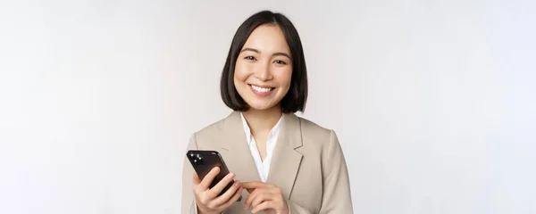Retrato de cerca de la mujer coreana, dama corporativa en traje, usando el teléfono móvil y sonriendo, sosteniendo el teléfono inteligente, de pie sobre fondo blanco — Foto de Stock