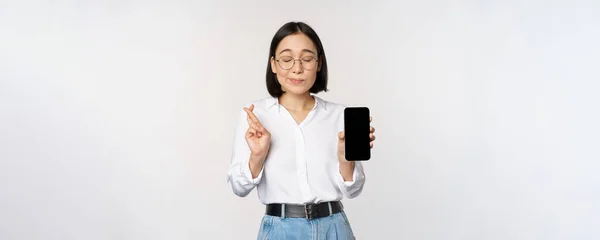 Hopeful young asian woman, showing mobile phone screen, app interface and fingers crossed, hoping for smth, making wish or paying, standing over white background — Stock Photo, Image