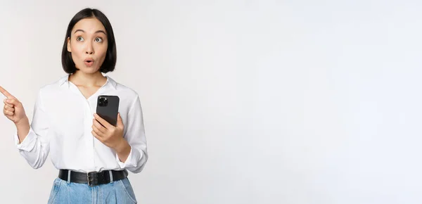 Возбужденная азиатская девушка делает покупки в мобильном приложении, держа телефон и указывая пальцем на пустое пространство для копирования, показывая промо-текст, стоя со смартфоном на белом фоне — стоковое фото