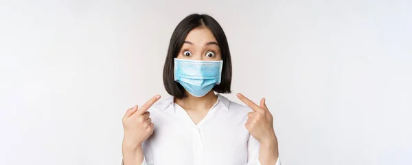 Imagen de la joven mujer asiática asombrada señalándose a sí misma mientras usa mascarilla médica, de pie sobre fondo blanco —  Fotos de Stock