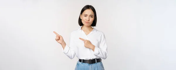 Porträt einer skeptischen asiatischen Frau, die die Stirn runzelt, mit dem Finger nach links zeigt und sich beschwert, Abneigung oder Missbilligung ausdrückt, vor weißem Hintergrund steht — Stockfoto