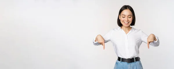 Retrato de mujer asiática feliz señalando con los dedos hacia abajo y mirando abajo en el anuncio, mostrando banner de información, publicidad, de pie sobre fondo blanco —  Fotos de Stock