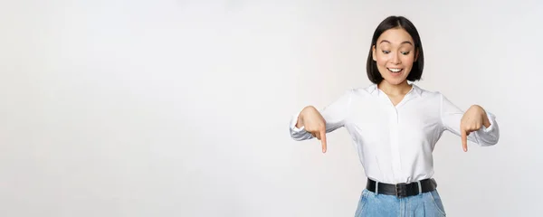 Portrét šťastné asijské ženy ukazuje prsty dolů a dívá se níže na reklamu, ukazuje info banner, reklama, stojící nad bílým pozadím — Stock fotografie
