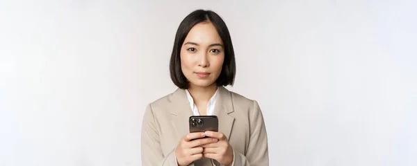 Immagine di donna d'affari asiatica in giacca e cravatta, in possesso di telefono cellulare, utilizzando l'app per smartphone, sorridente alla fotocamera, sfondo bianco — Foto Stock