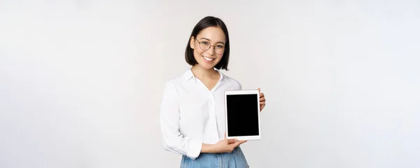 Young asian woman professional demonstrates digital tablet screen, info on her gadget, smiling and looking at camera, standing over white background — Stock Photo, Image