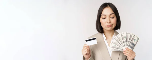 Pengusaha wanita yang bijaksana, korea perusahaan wanita menunjukkan kartu kredit dan uang tunai, dolar di tangan, berdiri di atas latar belakang putih dan berpikir — Stok Foto