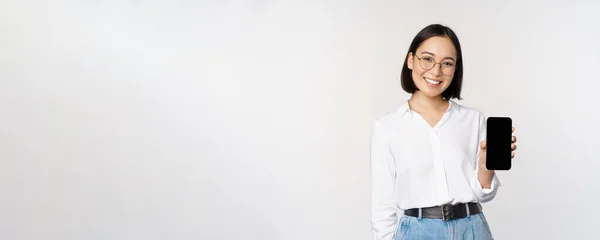 Mujer coreana sonriente mostrando la pantalla del teléfono inteligente, demostrando aplicación móvil, de pie sobre fondo blanco —  Fotos de Stock